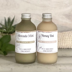 May include: Two glass bottles with silver lids and labels. The bottles are filled with a light brown liquid. The labels read "Avocado Mint" and "Honey Oat" with the text "Shine | Strength | Health" and "A.C.V. Hair Rinse Concentrate" on each.