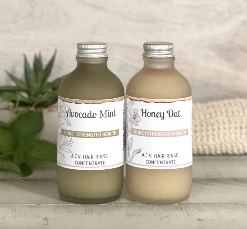 May include: Two glass bottles with silver lids and labels. The bottles are filled with a light brown liquid. The labels read "Avocado Mint" and "Honey Oat" with the text "Shine | Strength | Health" and "A.C.V. Hair Rinse Concentrate" on each.