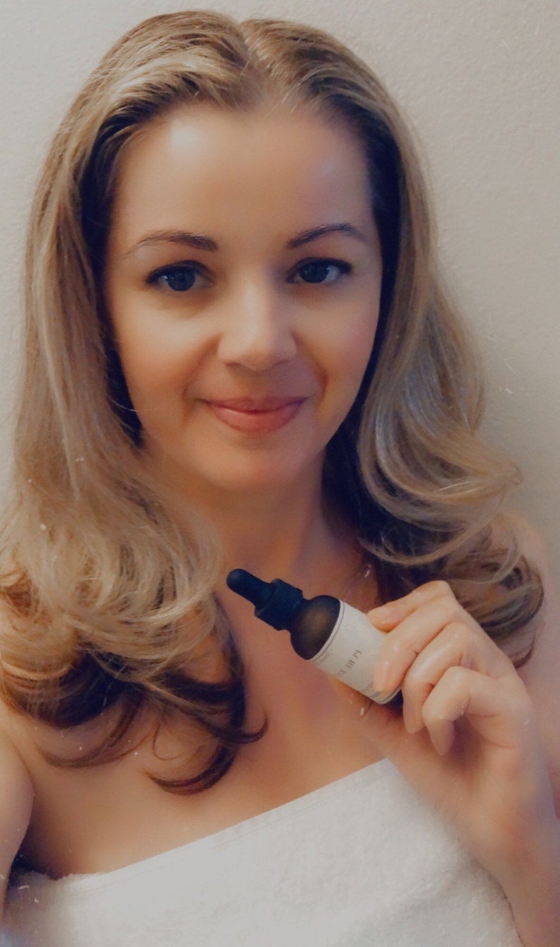 May include: A woman with long blonde hair is holding a small, dark glass bottle with a dropper. The bottle has a white label with the word "LUMI" printed on it. The woman is wearing a white towel.