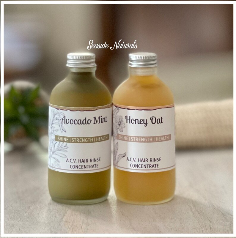 May include: Two glass bottles with silver lids and labels. The bottles contain liquid. The labels read "Avocado Mint" and "Honey Oat" with the text "Shine | Strength | Health" and "A.C.V. Hair Rinse Concentrate".