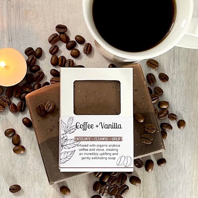May include: A brown and white bar of soap with a label that reads "Coffee + Vanilla" and "Exfoliate - Cleanse - Uplift". The soap is surrounded by coffee beans and a cup of coffee.