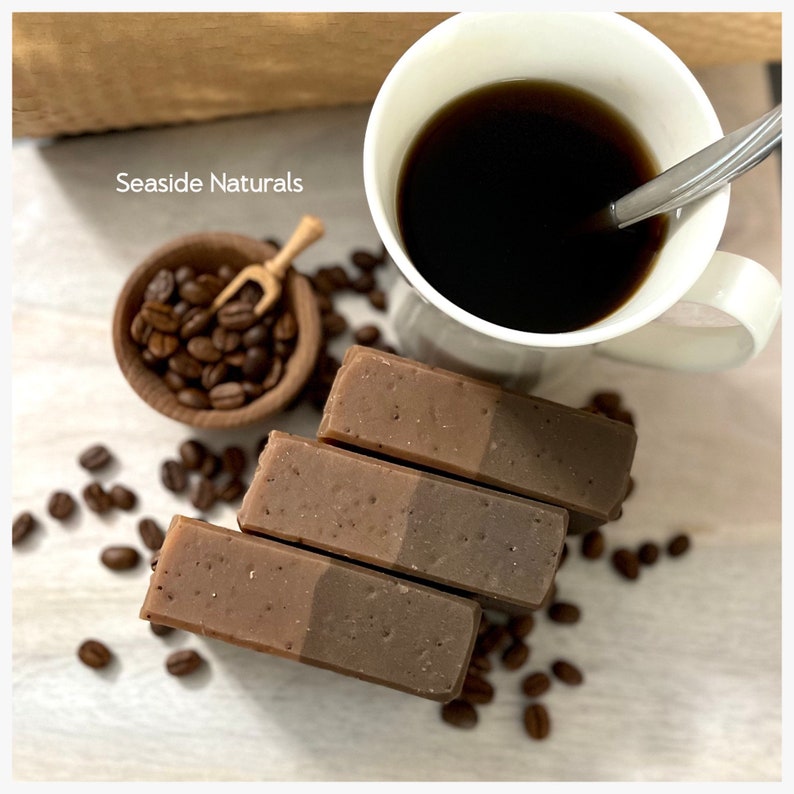 May include: Three bars of layered brown soap with coffee beans scattered around them. A cup of coffee and a small bowl of coffee beans are also in the image. The text "Seaside Naturals" is visible in the top left corner of the image.