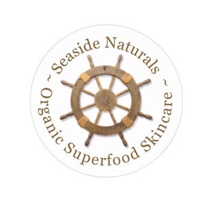 Puede incluir: Una pegatina circular blanca con texto marr&oacute;n que dice "~ Seaside Naturals ~ Organic Superfood Skincare". En el centro de la pegatina hay un tim&oacute;n de barco de madera marr&oacute;n.