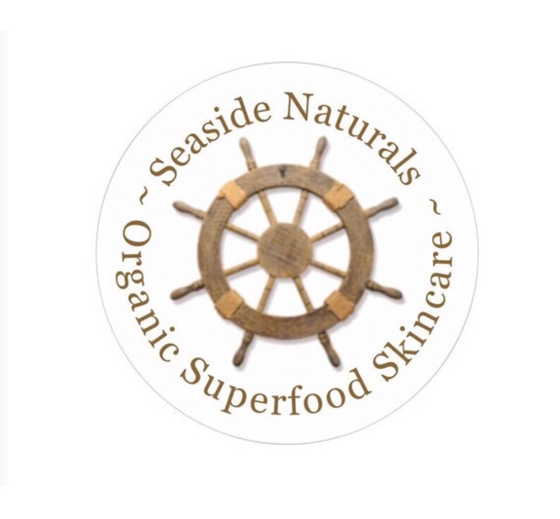 Puede incluir: Una pegatina circular blanca con texto marr&oacute;n que dice "~ Seaside Naturals ~ Organic Superfood Skincare". En el centro de la pegatina hay un tim&oacute;n de barco de madera marr&oacute;n.