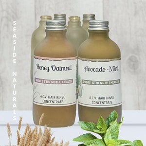 May include: Two amber glass bottles with silver lids and labels. The bottle on the left is labelled "Honey Oatmeal" and the bottle on the right is labelled "Avocado-Mint". Both bottles have the text "Shine | Strength | Health" and "A.C.V. Hair Rinse Concentrate" on their labels.