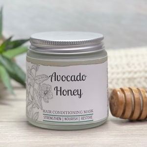 May include: A glass jar with a silver lid, labeled "Avocado Honey Hair Conditioning Mask." The label features botanical illustrations and the words "Strengthen, Nourish, Restore." A wooden honey dipper rests beside the jar, suggesting a natural hair care product.
