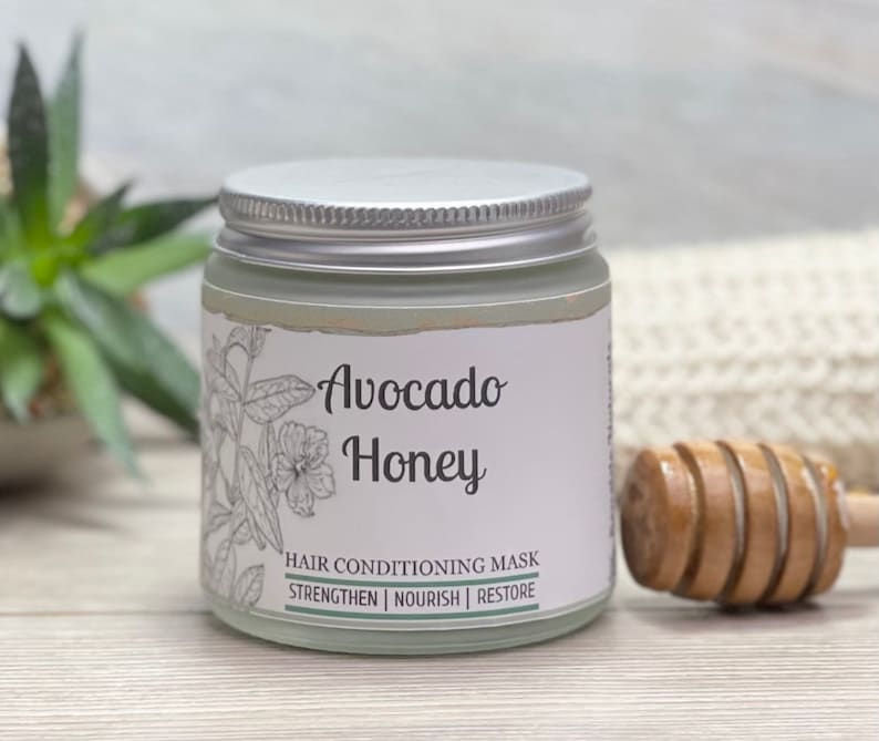 May include: A glass jar with a silver lid, labeled "Avocado Honey Hair Conditioning Mask." The label features botanical illustrations and the words "Strengthen, Nourish, Restore." A wooden honey dipper rests beside the jar, suggesting a natural hair care product.