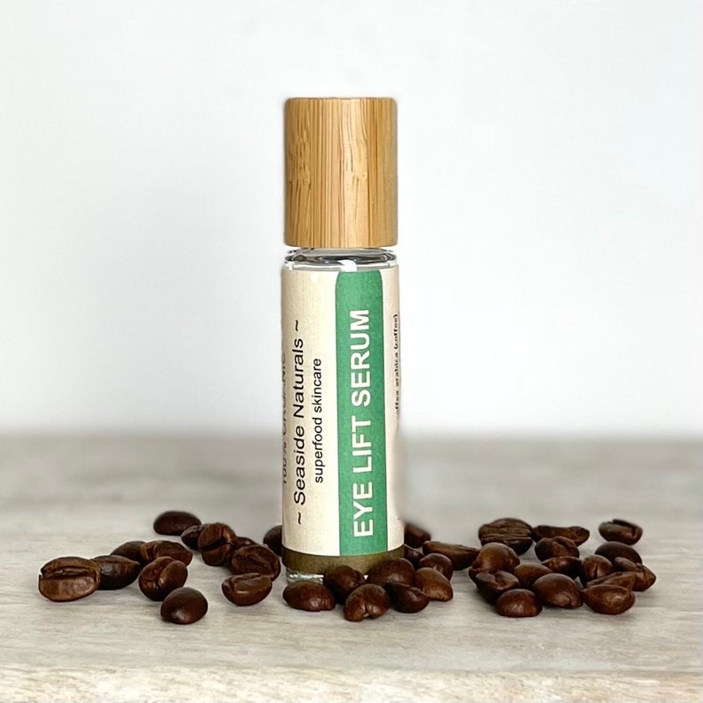 Puede incluir: Un peque&ntilde;o frasco de vidrio con tapa de bamb&uacute; y una etiqueta verde que dice "Eye Lift Serum - Seaside Naturals - superfood skincare". El frasco est&aacute; rodeado de granos de caf&eacute; sobre una superficie de madera clara.