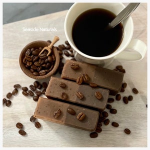 May include: Four bars of coffee-scented soap with coffee beans scattered around them. A cup of coffee and a small wooden bowl filled with coffee beans are also in the image. The text "Seaside Naturals" is visible in the top left corner of the image.