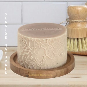 May include: A round, light brown soap bar with a delicate lace pattern on the side. The soap is sitting on a wooden soap dish with the words "Seaside Naturals" engraved on the bottom.