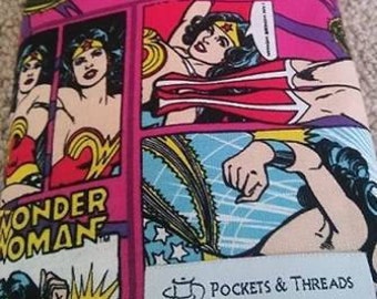 Puede incluir: Un artículo de tela enrollado con un diseño de cómic de Wonder Woman. La tela es predominantemente rosa, azul y amarillo, con detalles en negro. Una cinta negra está atada alrededor del rollo. El texto "Wonder Woman" y "Pockets & Threads" son visibles.