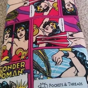 May include: A rolled-up fabric item featuring a Wonder Woman comic strip design. The fabric is predominantly pink, blue, and yellow, with black accents. A black ribbon is tied around the roll. The text "Wonder Woman" and "Pockets & Threads" are visible.