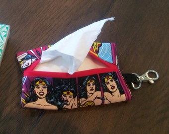 Puede incluir: Un porta pañuelos con temática de Wonder Woman con un borde rojo y una correa negra con un clip plateado. El porta pañuelos está lleno de pañuelos blancos y tiene un diseño de cómic de Wonder Woman.