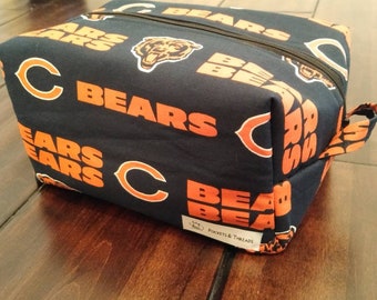 Puede incluir: Una bolsa de aseo azul marino con un estampado de logotipo de los Chicago Bears en naranja y blanco. La bolsa tiene una cremallera y un asa superior.