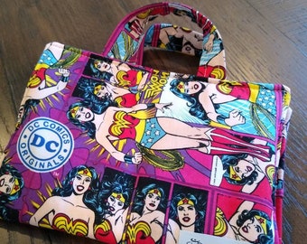 Puede incluir: Una bolsa de tela rosa con un estampado de cómic de Wonder Woman. La bolsa tiene un asa y una pequeña etiqueta que dice "Pockets & Threads".