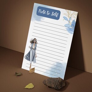 Può includere: Un blocco note bianco con linee nere orizzontali, con il testo Note to Self in un'intestazione blu. Il design include accenti acquerello blu, foglie in linea dorata e l'illustrazione di una persona che indossa un cappotto blu.
