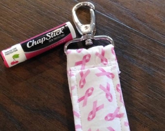 Puede incluir: Un llavero de tela rosa y blanco con un clip plateado y un patrón de cinta rosa. El llavero tiene un bolsillo para un tubo de bálsamo labial. El tubo de bálsamo labial es un ChapStick rosa y blanco con el texto "ChapStick Skin Protectant Cherry" impreso en el tubo.