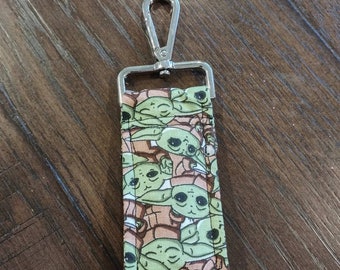 Puede incluir: Un llavero plateado con un charm de tela que presenta un patrón repetido de personajes de bebé Yoda verdes sobre un fondo marrón claro.