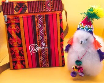 Puede incluir: Un bolso bandolera colorido con estampado, cierre de cremallera y correa marrón. Junto a él, una alpaca de peluche blanca con un sombrero festivo con borlas coloridas y un collar de cuentas. El bolso tiene la palabra "Peru" bordada.