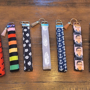 May include: A collection of key fobs with various patterns and designs. The key fobs feature patterns such as polka dots, anchors, paw prints, feathers, and a portrait. Each fob has a metal ring and a tassel.