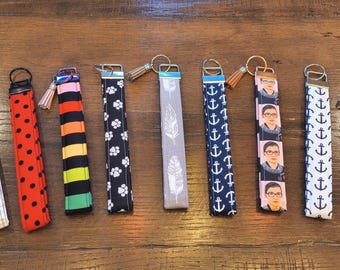Puede incluir: Una colección de llaveros con varios patrones y diseños. Los llaveros presentan patrones como lunares, anclas, huellas de patas, plumas y un retrato. Cada llavero tiene un anillo de metal y una borla.