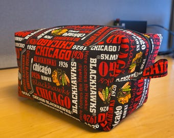 Puede incluir: Una bolsa rectangular negra y roja con cremallera de los Chicago Blackhawks. La bolsa está cubierta de texto y logotipos, incluyendo "Chicago Blackhawks 1926". La bolsa tiene una cremallera negra y un pequeño lazo de tela en el lateral.