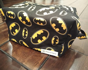 Puede incluir: Bolsa de aseo negra con un patrón repetido de logotipos de Batman amarillos. La bolsa tiene una cremallera y un asa. La bolsa tiene una etiqueta que dice "POCKETS & THREADS".
