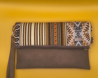 Puede incluir: Un bolso de mano marrón con una solapa superior estampada. La solapa presenta rayas verticales en tonos marrón, beige y blanco, con una sección de diseños geométricos. Se adjunta una correa de muñeca y una cadena plateada. El fondo es amarillo.