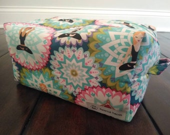 Puede incluir: Una bolsa de aseo rectangular de color verde azulado, rosa y verde con un patrón estampado, cierre de cremallera y asa de tela. La bolsa presenta un patrón repetitivo de flores estilizadas y figuras en poses de yoga. La bolsa tiene una etiqueta que dice "Pockets & Threads".