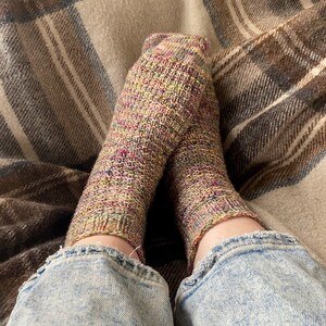 May include: A pair of hand-knitted socks in shades of beige, green, and pink, resting on a plaid blanket. The socks are worn with blue denim jeans, creating a cozy and comfortable aesthetic.
