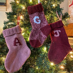 May include: Three knitted Christmas stockings in shades of red and pink hang on a Christmas tree. Each stocking has a letter: A, G, and J. The stockings are decorated with a ribbed cuff and a loop for hanging.
