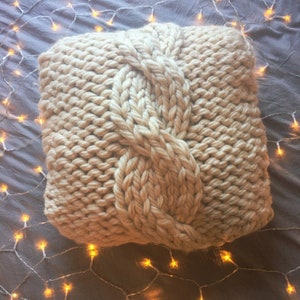 May include: A large, cream-colored knitted throw pillow with a cable knit pattern. The pillow is made of thick yarn and has a soft, cozy texture.