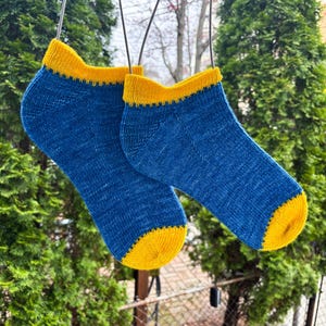 May include: A pair of blue ankle socks with yellow cuffs and toes hang from a wire. The socks are made of a knitted material and are displayed outdoors against a backdrop of green trees and a cloudy sky.