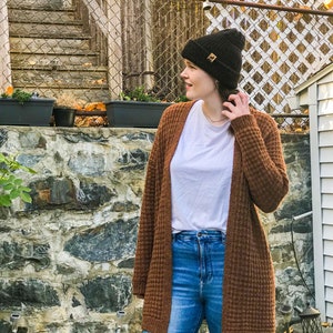 May include: A woman wearing a brown waffle knit cardigan, blue jeans, and a black beanie with a gold logo. She is standing in front of a stone wall with a wooden fence and fall foliage in the background.