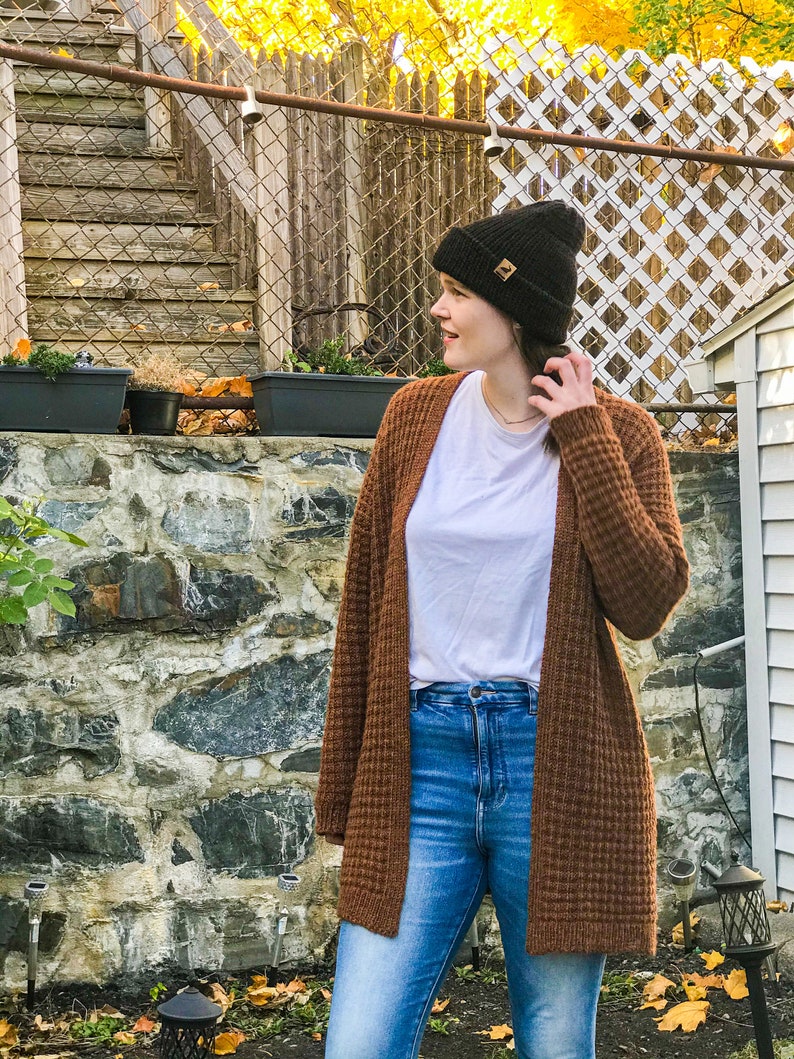 May include: A woman wearing a brown waffle knit cardigan, blue jeans, and a black beanie with a gold logo. She is standing in front of a stone wall with a wooden fence and fall foliage in the background.