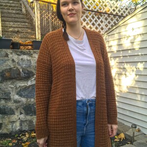 May include: A brown waffle knit cardigan with long sleeves and a relaxed fit. The cardigan is open in the front and is worn over a white t-shirt and blue jeans.