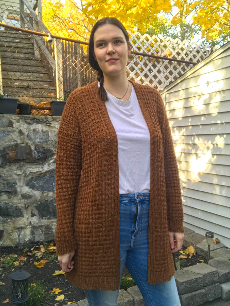 May include: A brown waffle knit cardigan with long sleeves and a relaxed fit. The cardigan is open in the front and is worn over a white t-shirt and blue jeans.