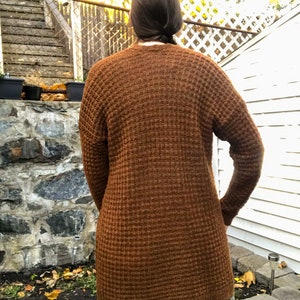 May include: A long brown waffle knit cardigan with long sleeves. The cardigan is made of a soft, comfortable material and has a relaxed fit.