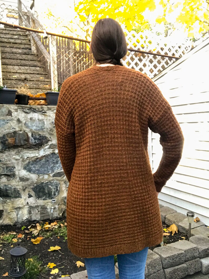 May include: A long brown waffle knit cardigan with long sleeves. The cardigan is made of a soft, comfortable material and has a relaxed fit.