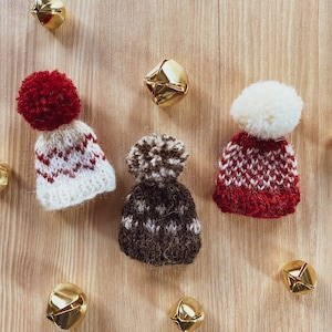 Peut inclure: Trois bonnets en tricot avec des pompons en rouge, marron et blanc avec un motif rouge et blanc. Les bonnets sont sur une surface en bois avec des clochettes dorées éparpillées autour d'eux.