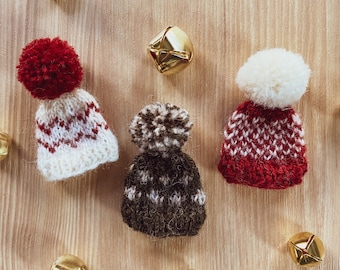 May include: Three knitted hats with pom-poms in red, brown and white with a red and white pattern. The hats are on a wooden surface with gold bells scattered around them.