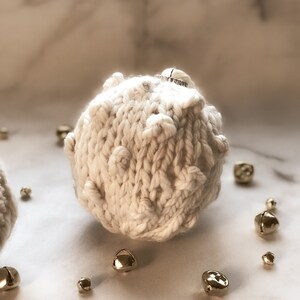 May include: A white knitted Christmas bauble with a small silver ring on top. The bauble is surrounded by small gold bells.