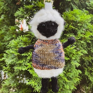 May include: A handcrafted sheep keyring with a black face, arms, and legs. The sheep is dressed in a colourful knitted sweater and a white fluffy hood. The keyring has a gold-coloured clasp.