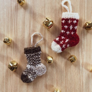 May include: Two knitted Christmas stockings, one red and white with a white snowflake pattern, and one brown and white with a checkered pattern, are displayed on a wooden surface with gold jingle bells scattered around them.