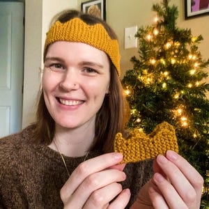 May include: Mustard yellow knitted crown headband and a matching small crown. The headband is worn on the head, and the small crown is held in the hand. The crowns have a wavy, pointed design. The background includes a Christmas tree and framed pictures.