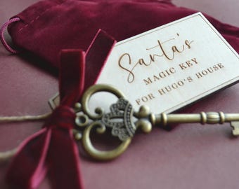 May include: An antique-style brass key with an ornate bow, tied with a deep red velvet ribbon to a rectangular wooden tag. The tag features the engraved text Santa's MAGIC KEY FOR HUGO'S HOUSE. A dark red velvet drawstring pouch is included.