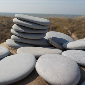 May include: A collection of smooth, grey stones, some stacked, others scattered. A coin is placed amongst the stones. The background shows a blurred view of the sea and dunes, creating a natural, minimalist aesthetic. The stones are approximately 10-15 cm in length.