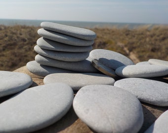 May include: A stack of smooth, gray stones balanced on a larger rock, with several other stones scattered around. A coin rests among the stones. The background features a blurred view of the sea and dunes, creating a natural, minimalist aesthetic.