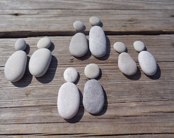 May include: Arrangement of smooth, gray stones on a weathered wooden surface. The stones are positioned to resemble family figures, with larger oval stones representing adults and smaller round stones as children. The natural light casts soft shadows.