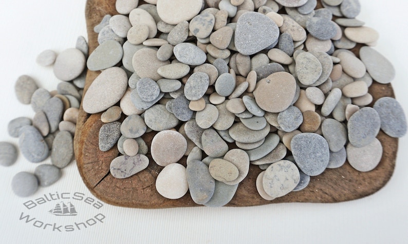 May include: A collection of smooth, gray and beige stones, some round and some oval, arranged on a wooden surface. The stones are of various sizes and are perfect for crafts or decoration.